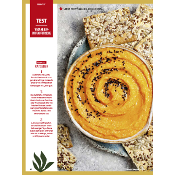 Das Bild zeigt eine Schüssel mit orangefarbenem Aufstrich, der mit schwarzen Sesamsamen bestreut ist. Um die Schüssel herum liegen mehrere knusprige Cracker mit verschiedenen Saaten. Der Hintergrund ist ein leicht strukturierter, heller Untergrund. Das Bi